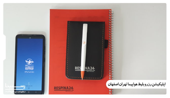 یک دفتر و دفترچه و خودکار و گوشی با صفحه اپلیکیشن رزرو بلیط هواپیما تهران اصفهان